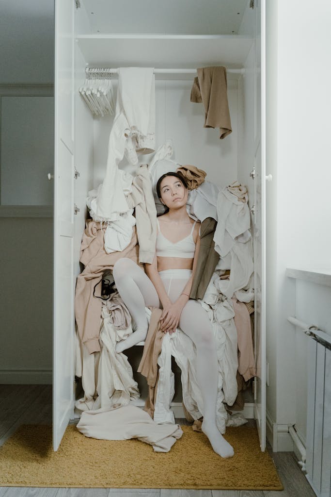 A contemplative woman sits surrounded by clothes in a messy wardrobe, evoking themes of consumerism and clutter.