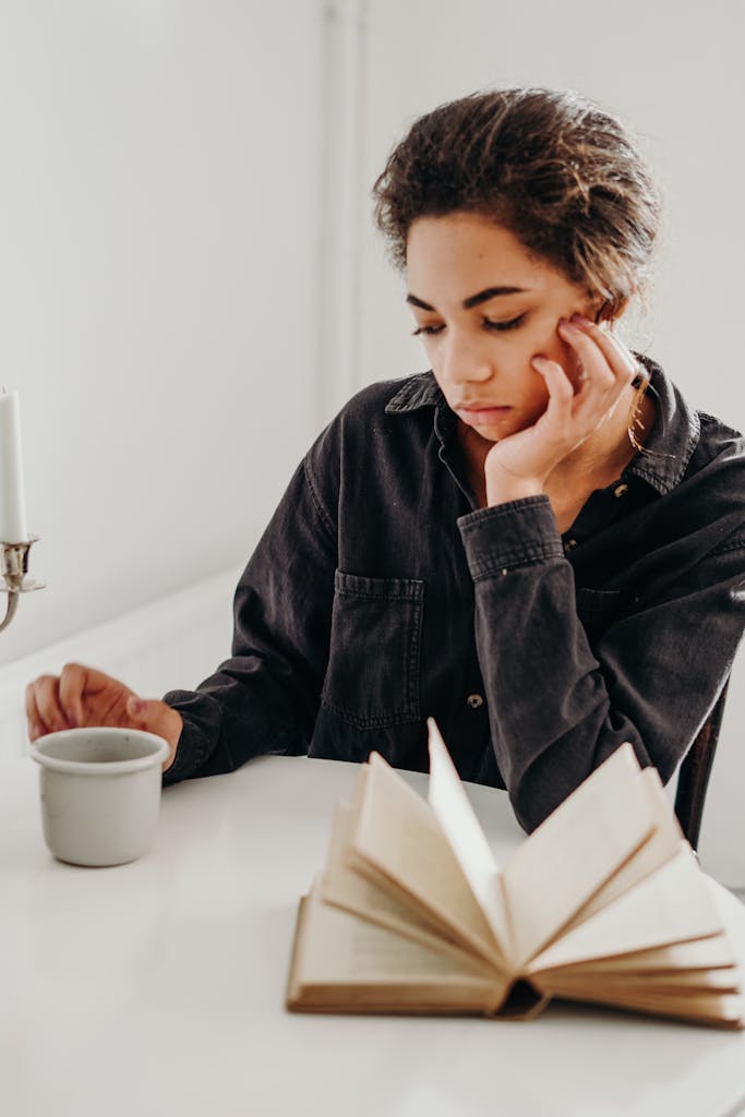 Pensive woman reading a book and enjoying coffee indoors, captured in a contemplative moment.