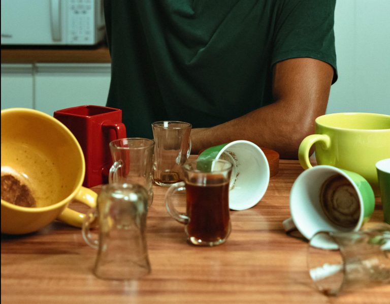 Young man drinking coffee straight from pot surrounded by mugs in kitchen.