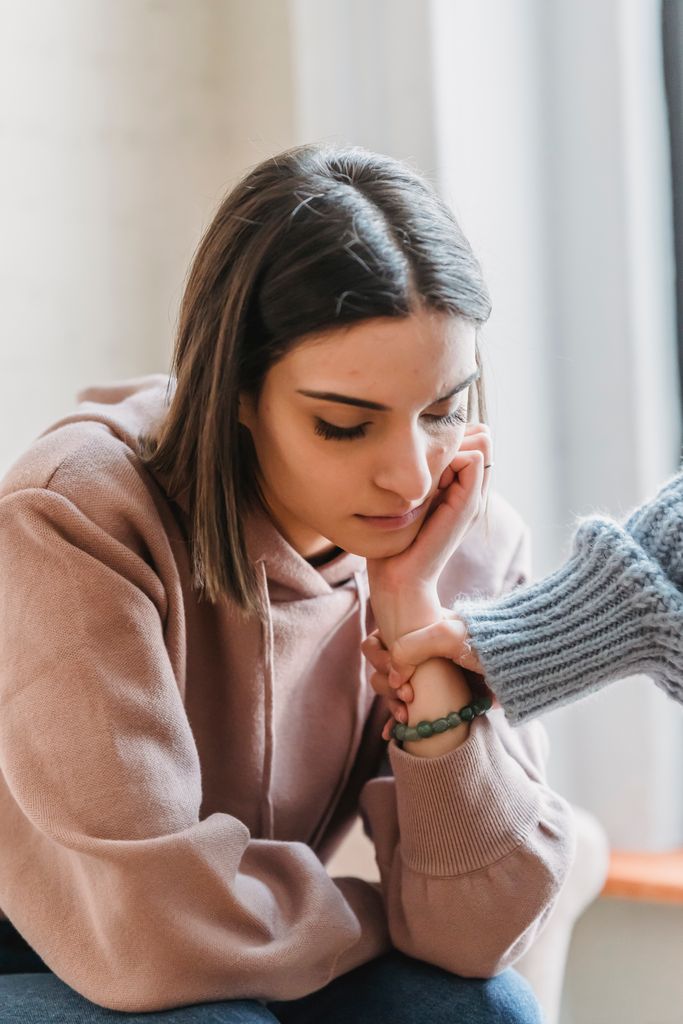 Faceless female grabbing hand of unhappy pensive girlfriend sitting in light living room and looking down during quarrel at home